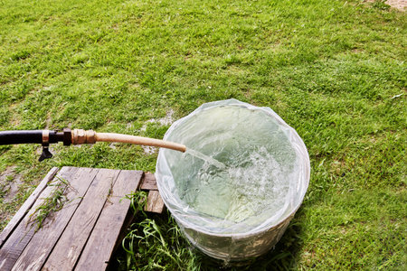 Guiding flow from hose into barrel liner, garden enthusiast fills rain barrel with water on lawn in...