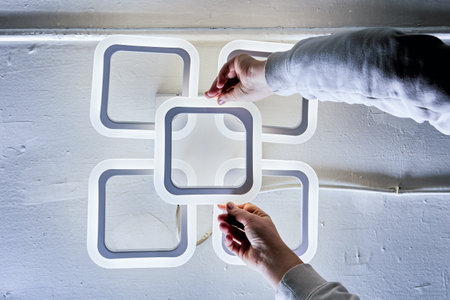 Modern LED fixture mounting process visible, square components arranged in cross pattern, white outer frames with gray inner details, human hands securing installation.の写真素材