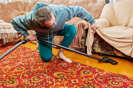 Cleaning rural house, homeowner uses vacuum clean dust floor before armchair, he stands one knee, lifting cover which covers armchair.の写真素材