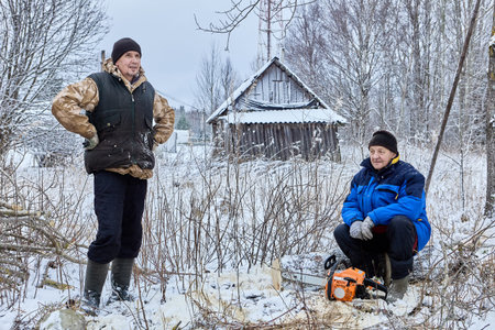 Worker crouches with chainsaw while fellow lumberjack stands nearby among bare trees and snow.の写真素材