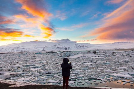 A woman in silhouette is phototraphing Glacier Lagoon (Jokulsarlon) at sunset with her mobile phone...