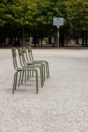A line of four green chairs on a concrete basketball court with the goal in the background with treesの写真素材