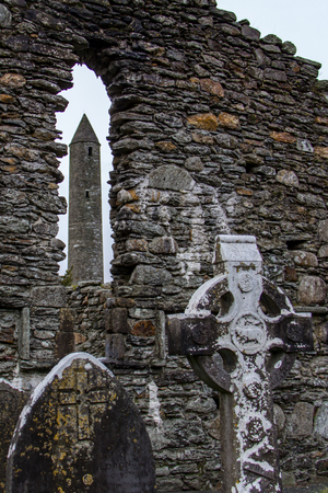 The Round Tower viewed through a window in a stone wall with a Celtic cross and grave stones in the foreground at the Glendalough Monastic Site in Wicklow, Irelandの写真素材