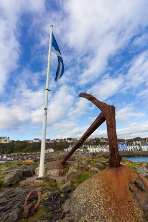 Scottish flag on a white flagpole and rusty boat anchor in Portpatrick harbor on an overcast day in Scotland, United Kingdomの写真素材