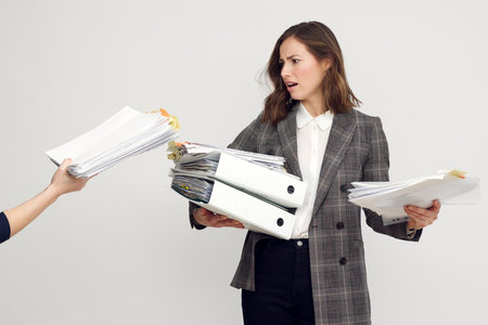 Stressed female worker and business woman holding a pile of paperwork while getting more work. Looking frustrated with too much work, isolated on white background.の写真素材