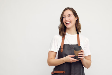 Portrait of beautiful young barista woman looking left and smiling or laughing, while holding a to-go coffee. Isolated on white background looking at copyspaceの写真素材
