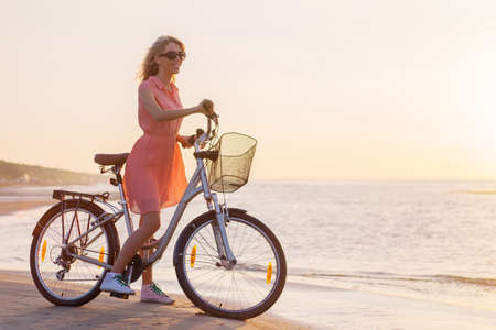 Young fashionable woman riding bicycle on the beach at sunsetの写真素材