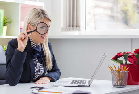 Business woman looking through a magnifying glass while sitting in front of computerの写真素材