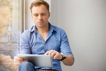 Young creative man sitting at window and using tablet computerの写真素材