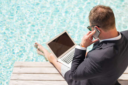 Businessman using computer and speaking on phone by the poolの写真素材