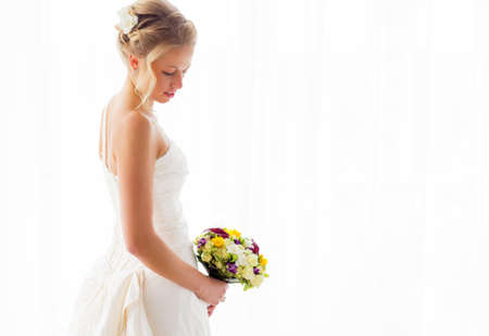 Bride standing by the window in her wedding gown and flowersの写真素材