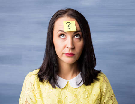 Woman with sticky note on her forehead looking up at question markの写真素材
