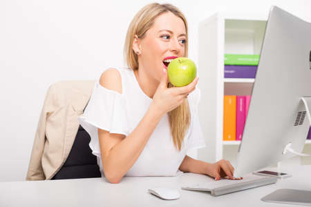 Woman eating fruit while working on computer in officeの写真素材