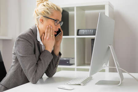 Woman talking on the phone and looking at computer in shockの写真素材