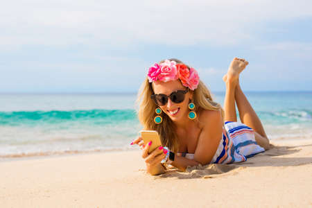 Happy young woman lying on the beach in sand and using mobile phoneの写真素材