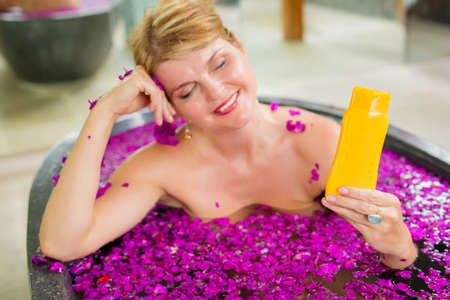 Woman showing sample beauty product bottle while sitting in bathの写真素材