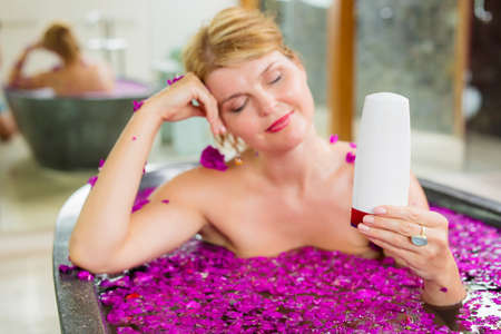 Woman holding bod care product bottle in hand while relaxing in bathの写真素材