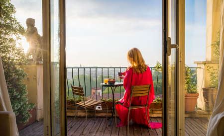 Woman sitting on balcony and overlooking city in morning at sunriseの写真素材