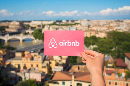 Rome, Italy - May 13, 2018: Person holding Airbnb logo in hand with city in background.のeditorial素材
