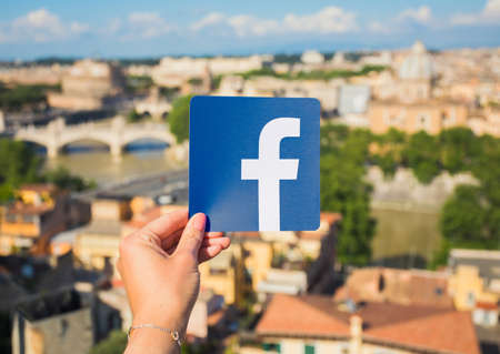 Rome, Italy - May 13, 2018: Person holding Facebook logo in hand with city in background.のeditorial素材