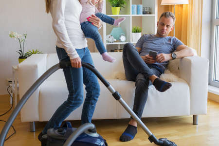 Woman with small child doing housekeeping while man sitting in couchの写真素材