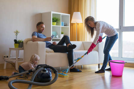 Woman cleaning floors at home while her lazy man sitting in couchの写真素材