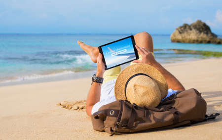 Man buying airline tickets online on tablet while relaxing on beachの写真素材