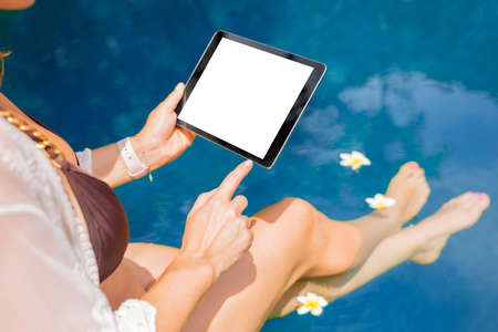 Woman sitting by the pool and using tablet computer. Horizontal white screen.の写真素材