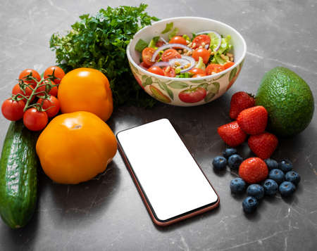 Smartphone with white empty screen  on a kitchen table surrounded by vegetables and fruitsの写真素材