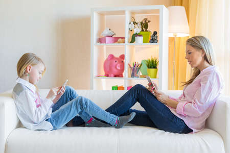 Mother and daughter using phones while sitting on sofa. Smartphone  addiction concept.の写真素材