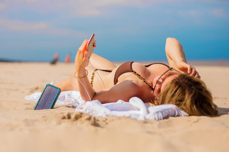 Woman charging mobile phone with portable solar power battery bank while lying on the beachの写真素材