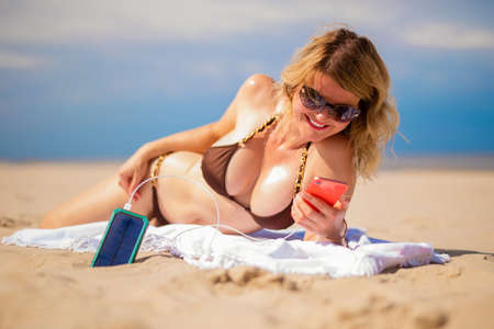 Woman using solar power bank to charge her mobile phone while relaxing on the beachの写真素材