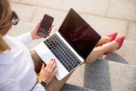 Woman using laptop and mobile phone while sitting outsideの写真素材