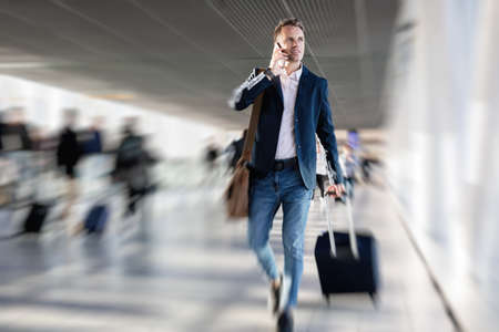 Man walking in airport carrying hand luggage while talking on phoneの写真素材