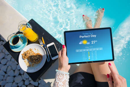 Woman eating breakfast by the pool and looking at weather app which shows that today is a perfect day.の写真素材