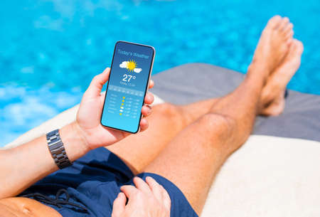Man relaxing by the pool in hot summer day and checking weather forecast on his mobile phone.の写真素材