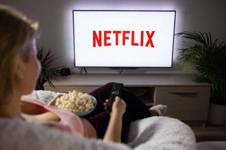 Riga, Latvia - April 14, 2021: Woman holding popcorn and watching TV at home with Netflix logo on the screen.のeditorial素材