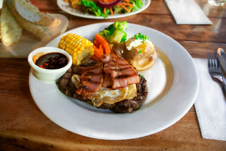 Plate of delicious piece of well done stake with fried egg and bacon on top. Serve with BBQ sauce, corn, green vegetable and bake potato. Garlic cheese bread on the sideの写真素材