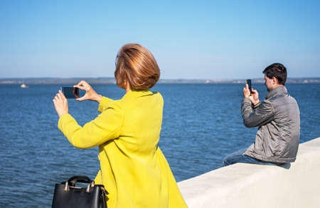 Young people are sitting on the seafront and holding smartphones in their hands. The concept of social distancing. Selective focus.の写真素材