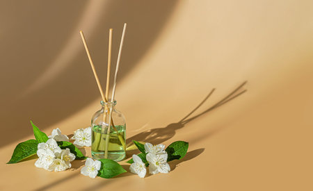 A glass bottle of aromatic oil with bamboo sticks and jasmine flowers on a beige surface. Copy...