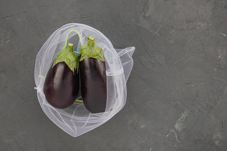 Two fresh purple eggplants placed in a reusable eco-friendly mesh bag on a dark gray textured surface. Perfect for concepts of sustainability, healthy eating, and zero waste lifestyle.の写真素材