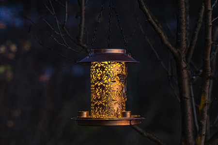 Illuminated bronze bird feeder lantern softly glows in the twilight of an outdoor garden. Cozy and inviting, perfect accent for wildlife-friendly backyard and peaceful night ambience.の写真素材