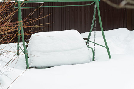 Empty green metal garden swing bench covered with a heavy layer of fresh white snow. Winter backyard scene with a brown fence and snowy nature.の写真素材