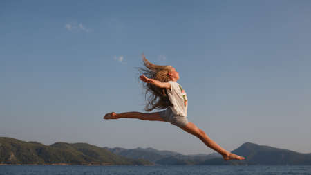 The jump, flight above the sea  and mountains of the young girl about 9-12 years old with the blonde and loose hair in the rays of the setting sun. She wears top-shirt and shorts.の写真素材