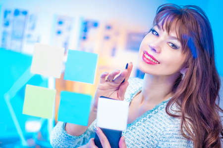 Business woman is engaged in planning. Colored stickers on the glass wall. The girl in the office is planning a meeting. Friendly smile. Brunette makes notes on colored stickers.の写真素材