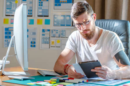 The designer compares the design of the site on the tablet. Website development for mobile devices. Graphic designer is developing a website for mobile devices. Remote work. Office graphic designer.の写真素材