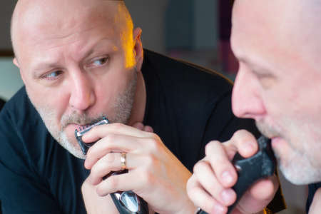 The man at the mirror is cutting his beard. A man shaves his beard. Beard trimming with a trimmer. Care of appearance. Trimmer for haircuts.の写真素材