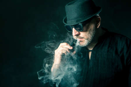 A man smokes electronic cigarettes. Aged man in smoke. Vape. Smoke from smoking. A man with a gray beard smokes.の写真素材