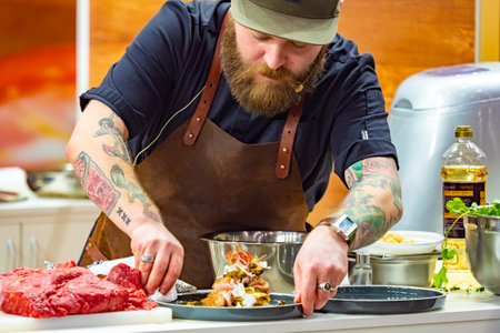 A man demonstrates the preparation of a meat dish. A cook with a beard and a leather apron. The men's restaurant kitchen. Serving meat dishes. The chef with the tattoo. 06.11.2019のeditorial素材