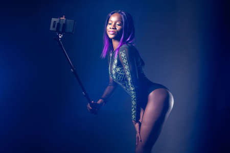Selfie girl. Black afro model takes a photo with a selfie stick. Girl in a black bodysuit posing. Selfshot. Portrait with a selfie stick. Photo for social networks. Egomania. Fashion trend.の写真素材